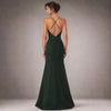 A back view of a model posing in a studio with a grey gradient background wearing a luxury emerald green satin evening gown by Cenia New York. The floor-length gown features an asymmetric neckline, a structured A-line sheath silhouette, and elegant hand-draped fabric cascading down the front, gathered at the waist with an intricate black beaded embellishment.