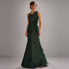 A model poses in a studio with a grey gradient background wearing a luxury emerald green satin evening gown by Cenia New York. The floor-length gown features an asymmetric neckline, a structured A-line sheath silhouette, and elegant hand-draped fabric cascading down the front, gathered at the waist with an intricate black beaded embellishment.