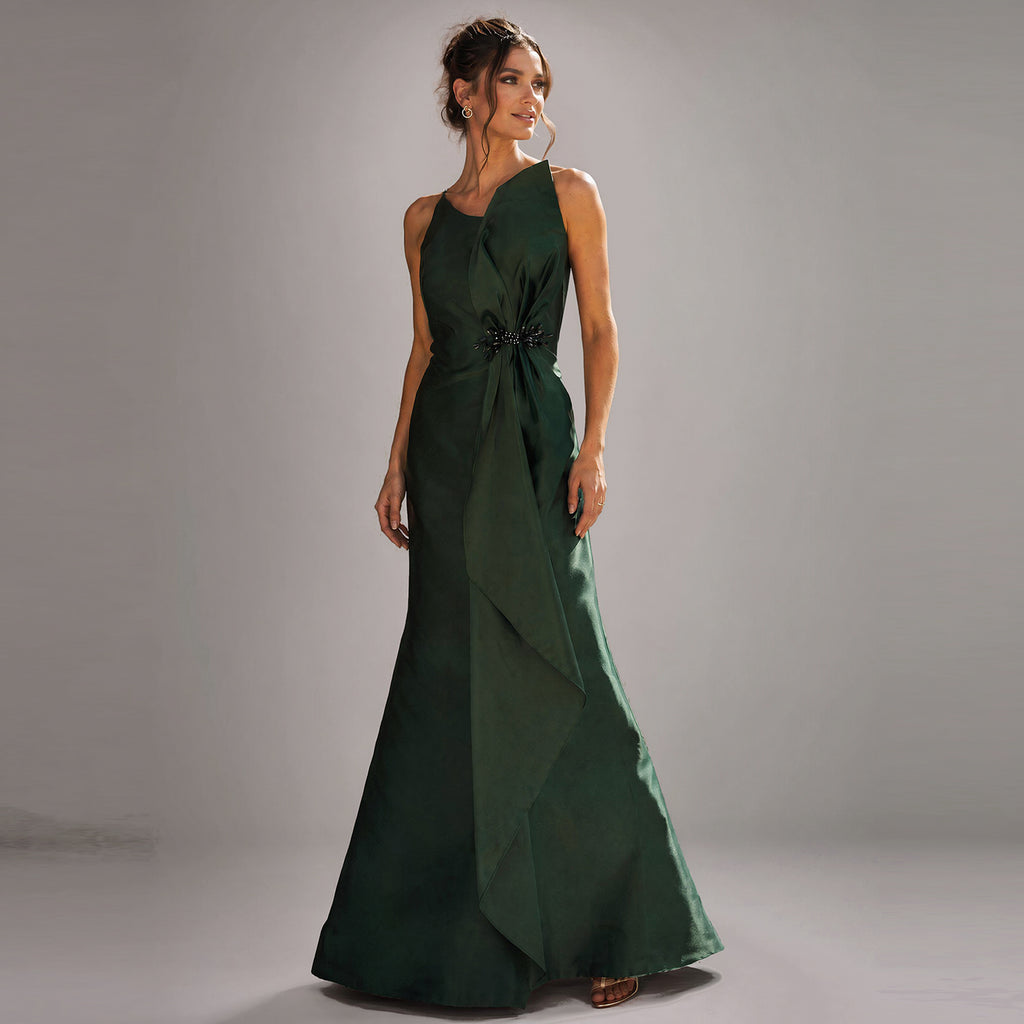 A model poses in a studio with a grey gradient background wearing a luxury emerald green satin evening gown by Cenia New York. The floor-length gown features an asymmetric neckline, a structured A-line sheath silhouette, and elegant hand-draped fabric cascading down the front, gathered at the waist with an intricate black beaded embellishment.