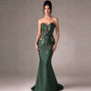 A woman models a strapless sweetheart mermaid gown in dark emerald green with gold floral embroidery, set against a neutral gray studio backdrop.