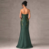 A woman stands with her back to the camera wearing a strapless emerald green mermaid gown with gold floral embroidery on the back, set against a textured gray studio backdrop.