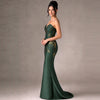 A woman models a strapless emerald green mermaid gown with gold floral embroidery, standing against a neutral gray studio backdrop.