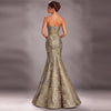 A woman shown from the back wearing a strapless mermaid-style evening gown with a fitted bodice, flared skirt, and ornate metallic floral pattern. The dress appears in a champagne-gold tone with a gold waistband detail, photographed against a smooth gray studio backdrop.