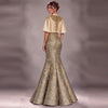 A woman modeling a floor-length evening gown from the back, featuring a champagne gold satin cape-style bodice with gold embroidery and a matching patterned mermaid skirt against a gray studio backdrop.