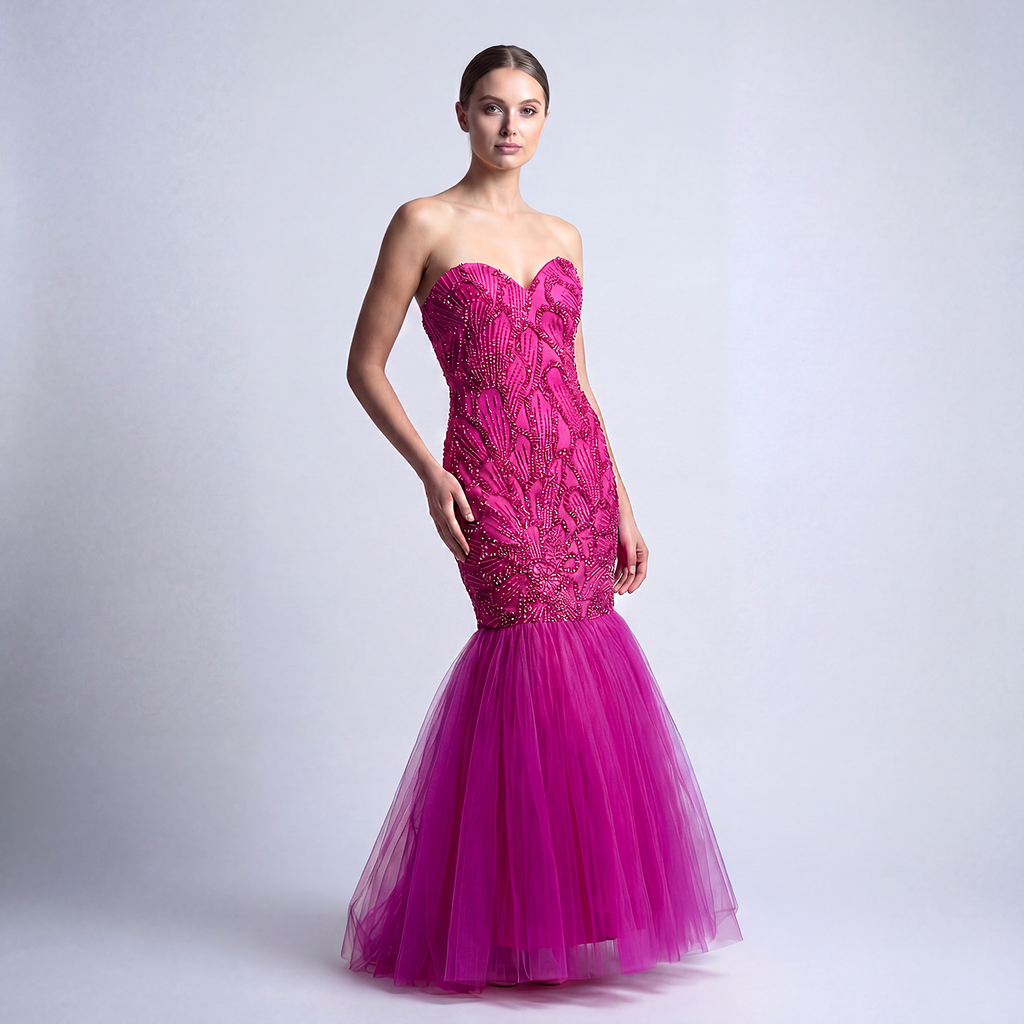 A woman wearing a bright fuchsia pink strapless mermaid gown with a heavily beaded bodice and a voluminous floor-length tulle skirt, standing against a neutral studio background.