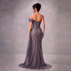 A woman stands with her back to the camera wearing a floor-length off-the-shoulder evening gown in a shimmering taupe-gray fabric, set against a textured gray studio backdrop.
