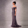 A woman models an off-the-shoulder, floor-length evening gown in a shimmering dark gray or charcoal purple fabric, standing against a textured gray studio backdrop.