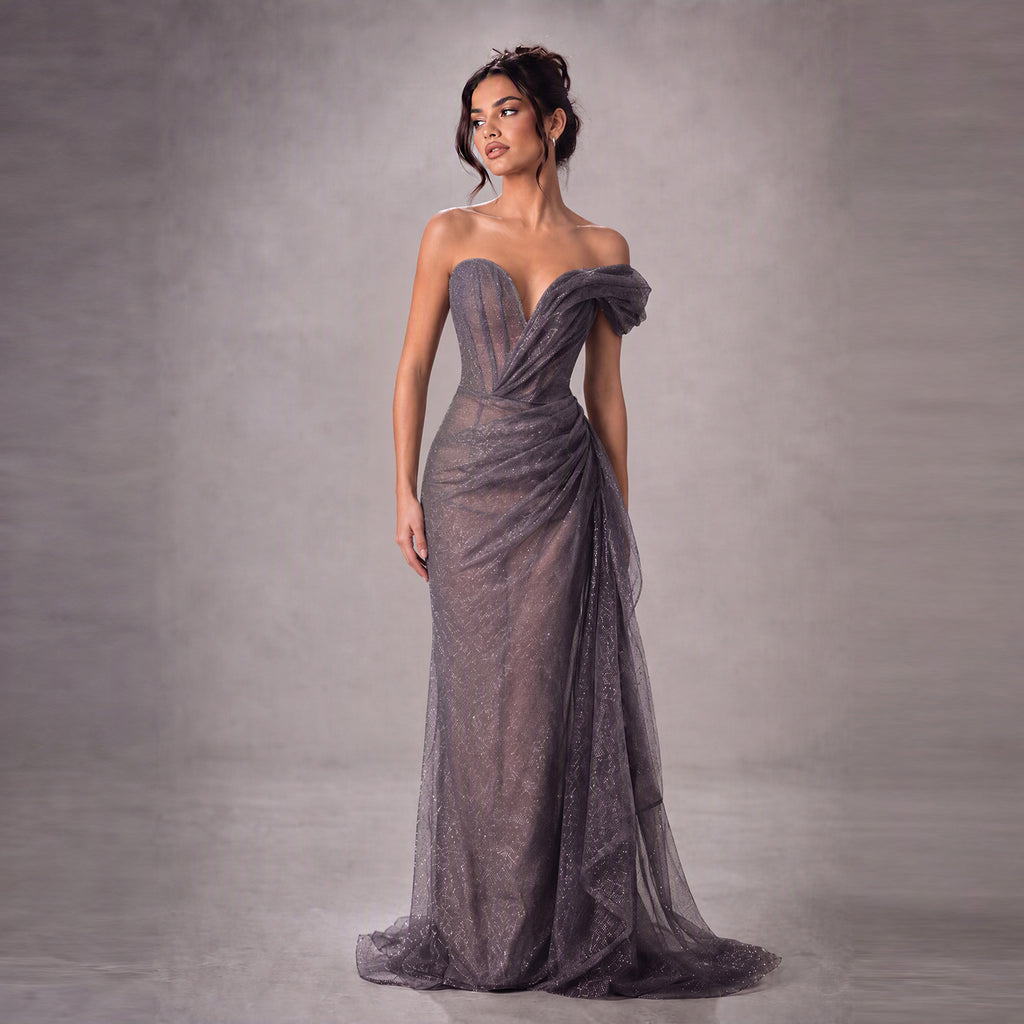 A woman models a strapless floor-length gown in a studio setting. The dress has a structured sweetheart bodice, draped off-shoulder ruffle sleeves, and a sheer textured tulle skirt with a train.