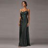 A woman modeling a floor-length dark green satin gown with thin spaghetti straps, a ruched sweetheart neckline, and 3D floral appliqués down the side, photographed against a solid gray studio background.