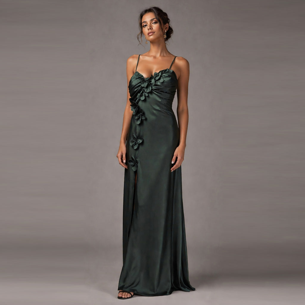 A woman modeling a floor-length dark green satin gown with thin spaghetti straps, a ruched sweetheart neckline, and 3D floral appliqués down the side, photographed against a solid gray studio background.