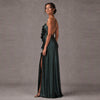 A woman modeling a floor-length dark green satin gown with a ruffled bodice, one-shoulder design, thin back straps, and a high slit, photographed against a neutral gray studio backdrop.