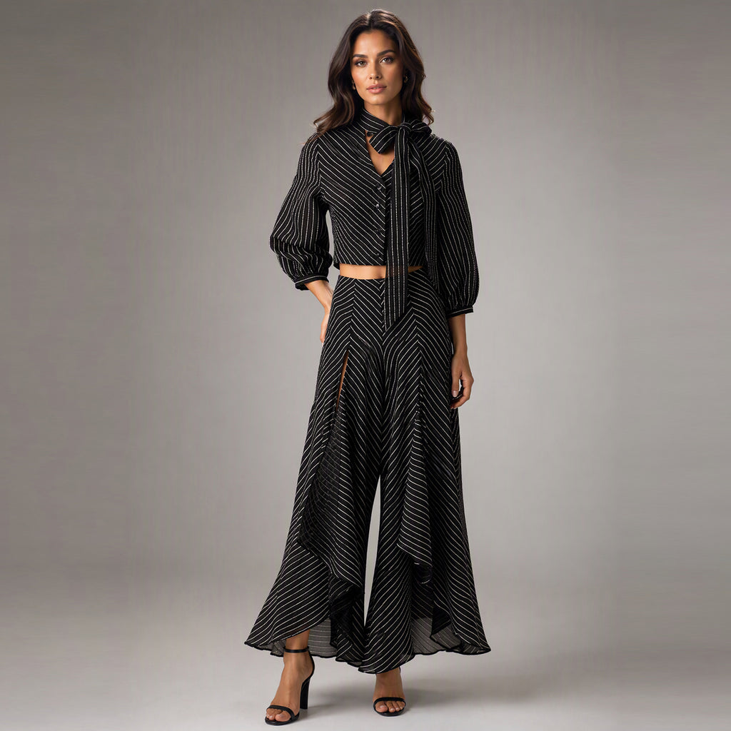 A woman modeling a black and white striped two-piece set consisting of a long-sleeve tie-neck blouse and matching high-low trousers, photographed against a neutral gray studio background.