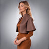 A woman modeling a brown suede-like cropped cape jacket over a rust-orange long-sleeve dress, photographed against a neutral gray studio background.